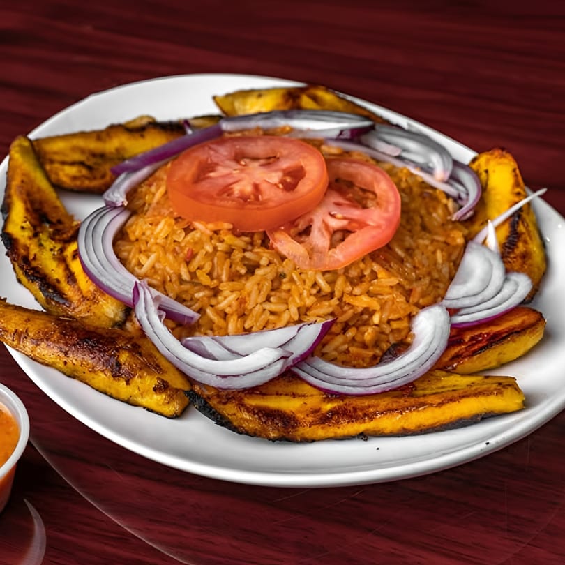 Jollof rice topped with sliced tomatoes and red onions, surrounded by fried plantains on a white plate.
