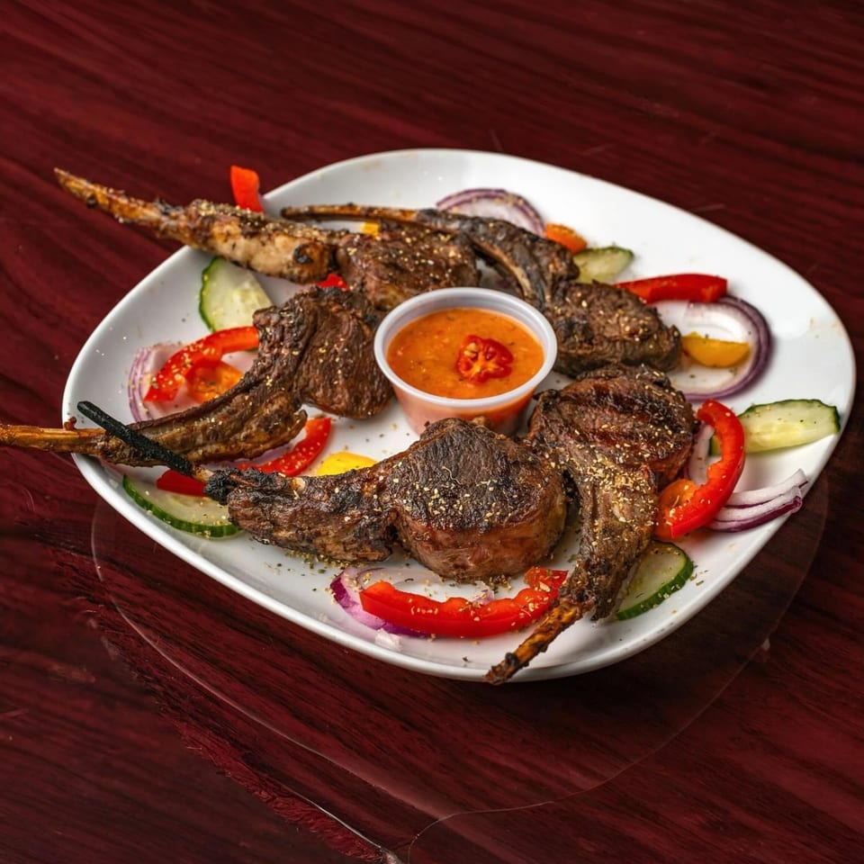 Grilled lamb chops, also known as Suya, with a side of dipping sauce and fresh vegetables.