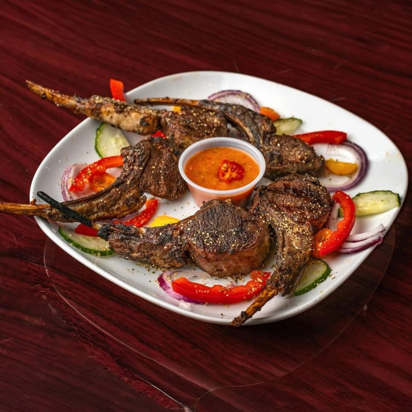 Grilled lamb chops, also known as Suya, with a side of dipping sauce and fresh vegetables.