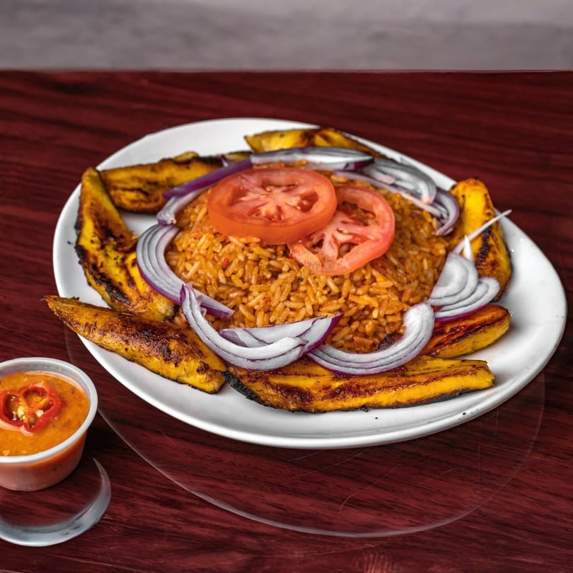 African Jollof rice with tomato slices, red onions, and fried plantains, served with a side of sauce.
